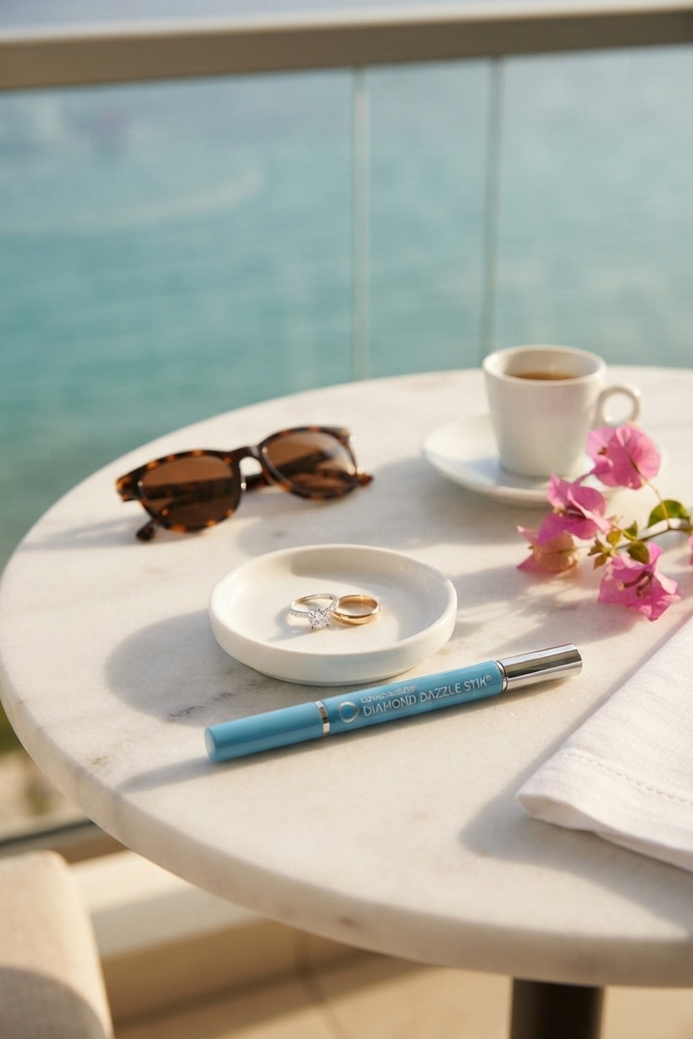Sky blue ring cleaner pen beside a white dish holding a diamond engagement ring and a gold band on a marble balcony table by the sea, with sunglasses and espresso